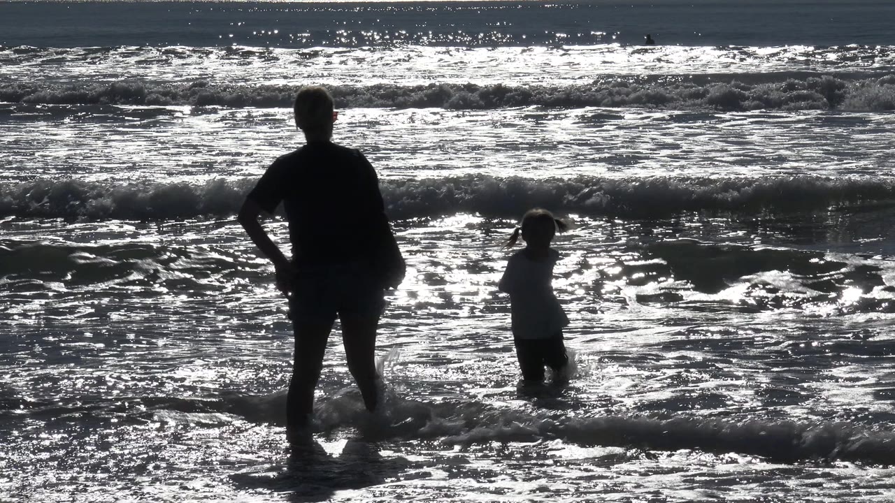 At Huntington Beach, California, the woman remains in silhouette while the child stands farther right in ankle-deep surf and a low wave breaks behind them.