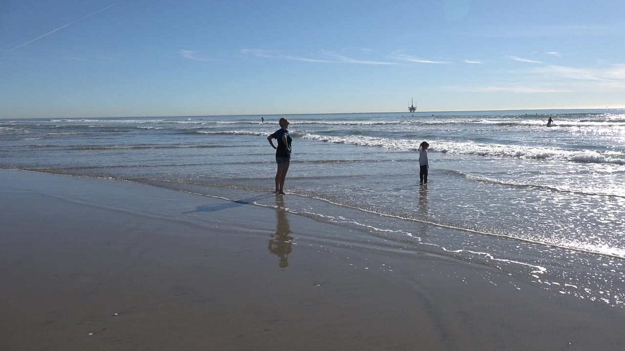 At Huntington Beach, an adult stands on wet sand while a child faces the incoming surf under a bright blue sky.