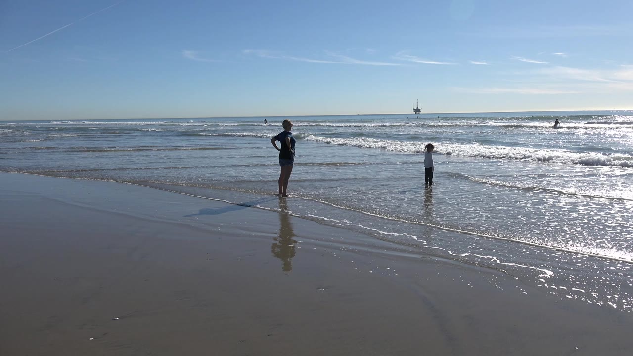At Huntington Beach, the child stands closer to a thin line of foam while the adult remains farther left on reflective wet sand.