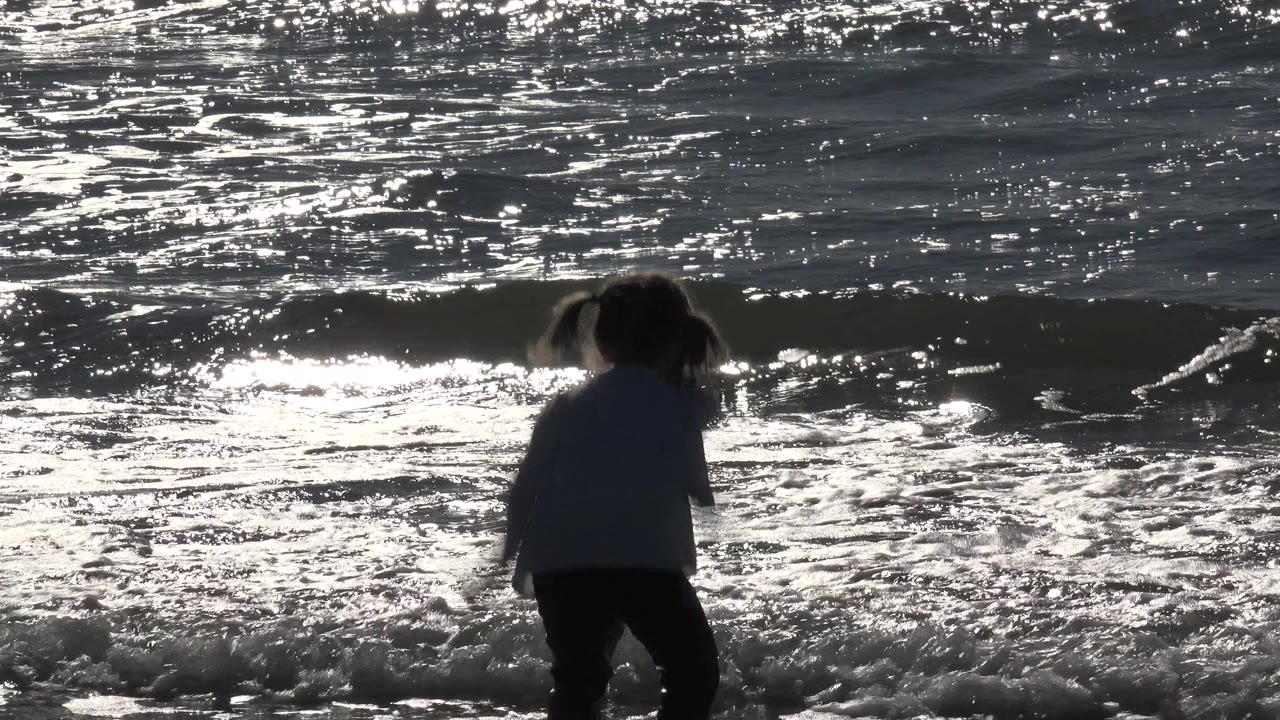 At Huntington Beach, California, a little girl stands in shallow surf as a small wave reaches her legs and sun glare covers the water behind her.