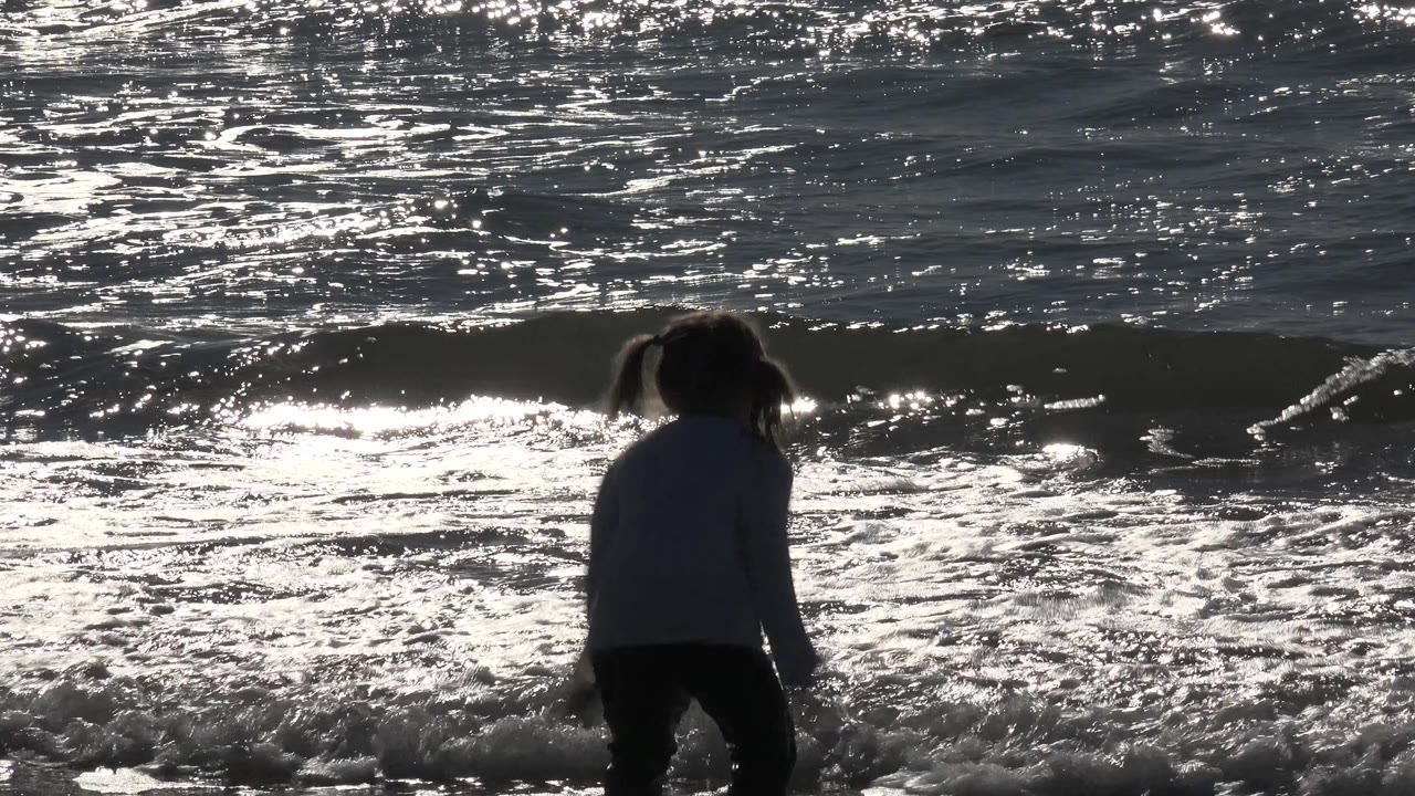 Backlit child crouching near a small wave in reflective ocean water.