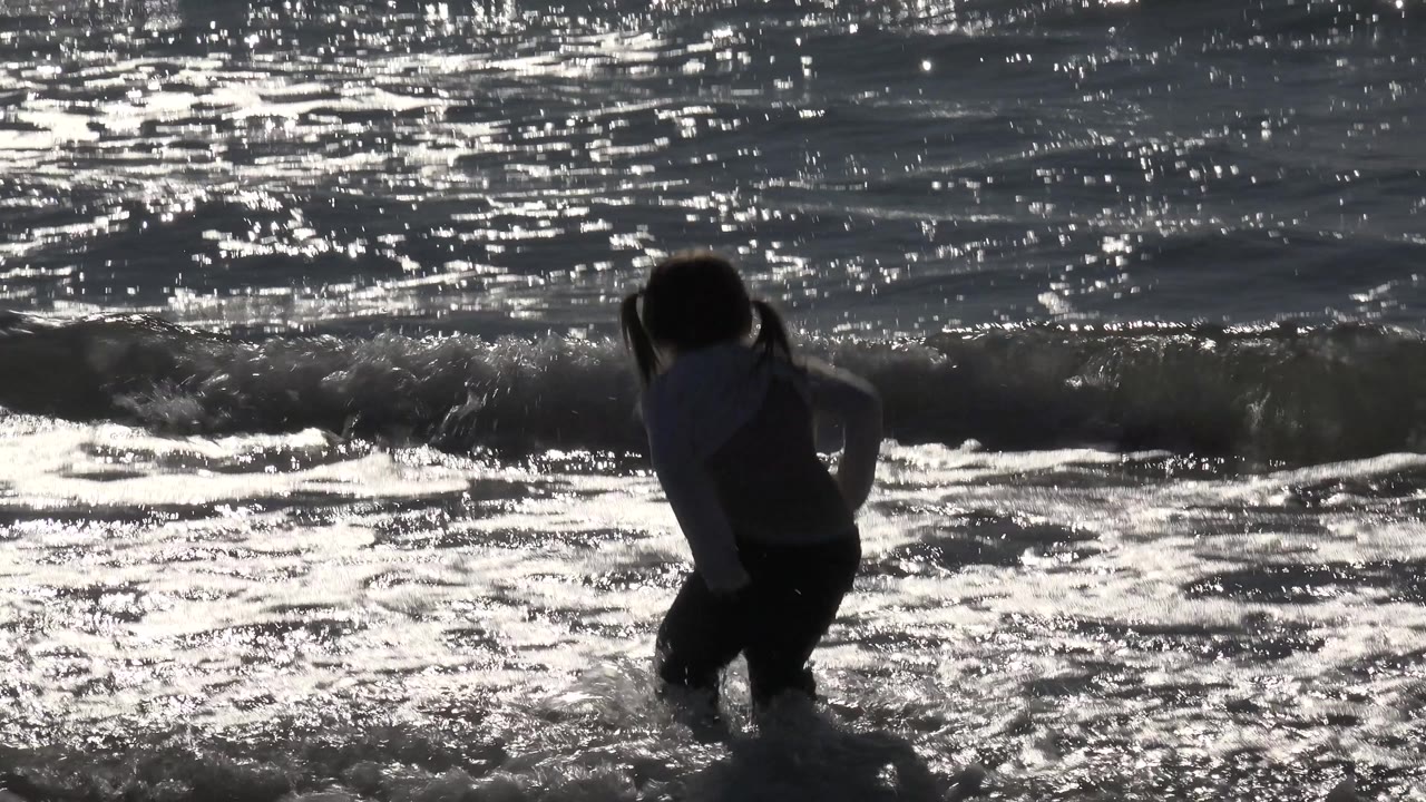 At Huntington Beach, California, the little girl lifts through the shore break as a small wave curls behind her and bright glare outlines her figure.