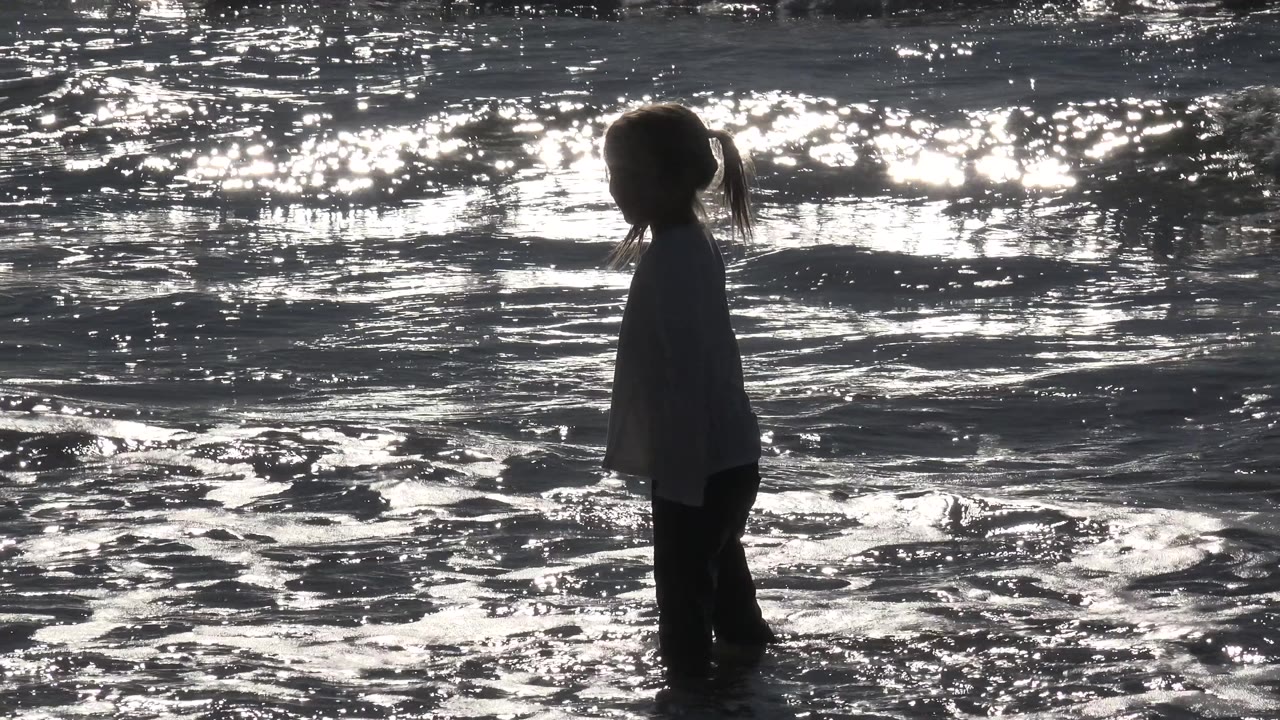 A young girl with pigtails stands side-on in shallow water at Huntington Beach, California, silhouetted against bright ocean reflections.