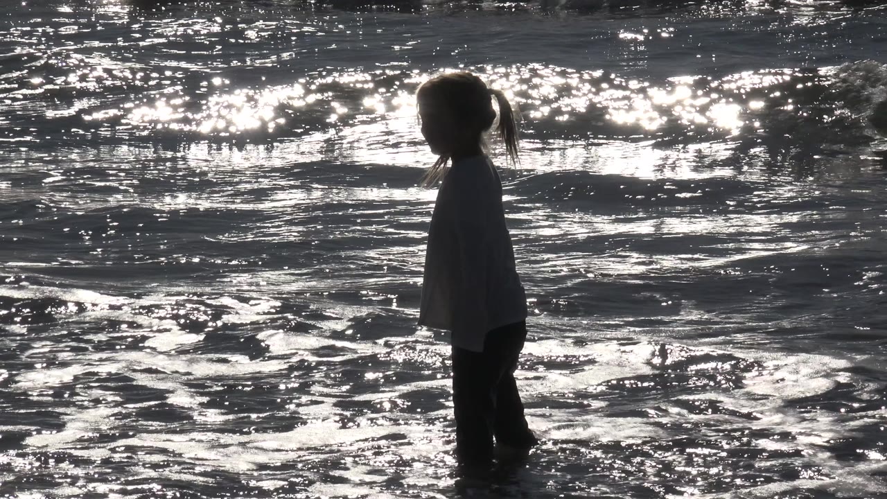 The child remains in profile at Huntington Beach, California, with rippled shallow surf and bright glare behind her.