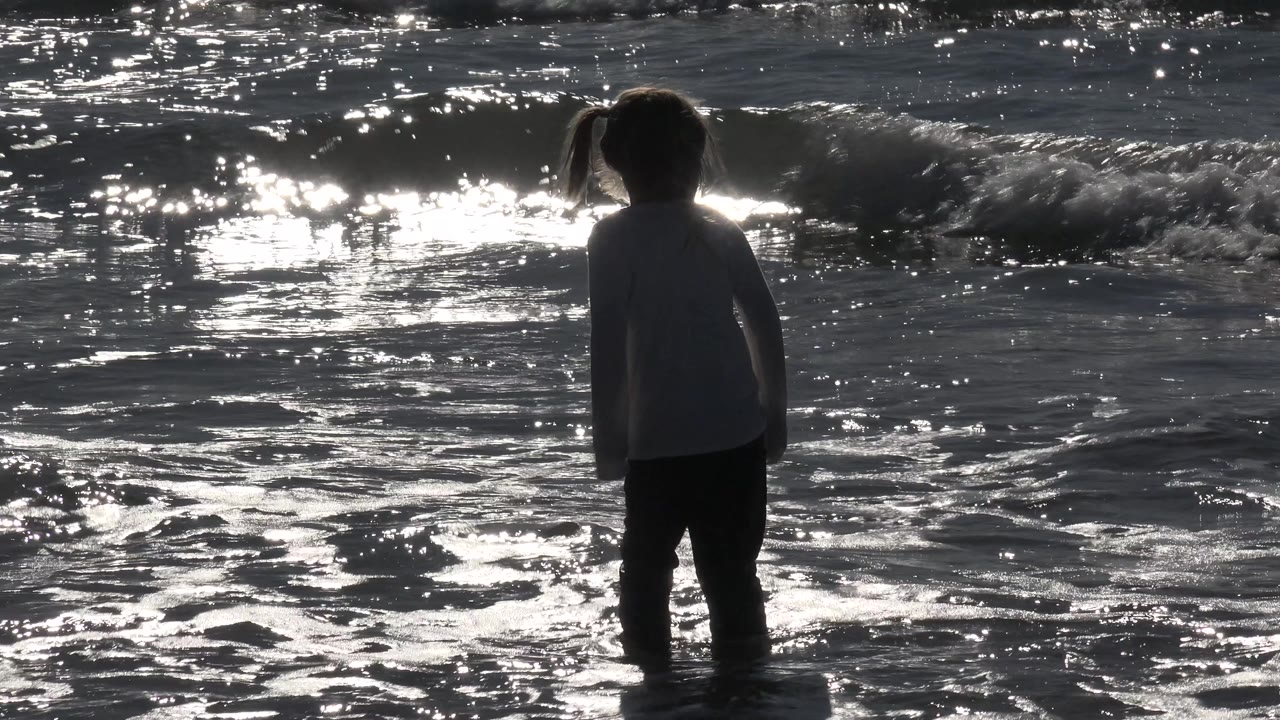 A young girl faces away toward the ocean at Huntington Beach, California, with a small wave line and bright reflections behind her.