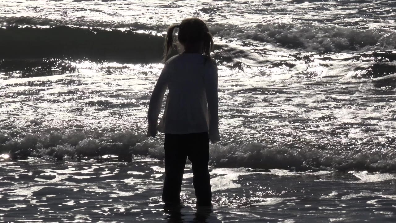 At Huntington Beach, California, a young girl stands ankle-deep in reflective surf while a foamy wave rolls behind her.