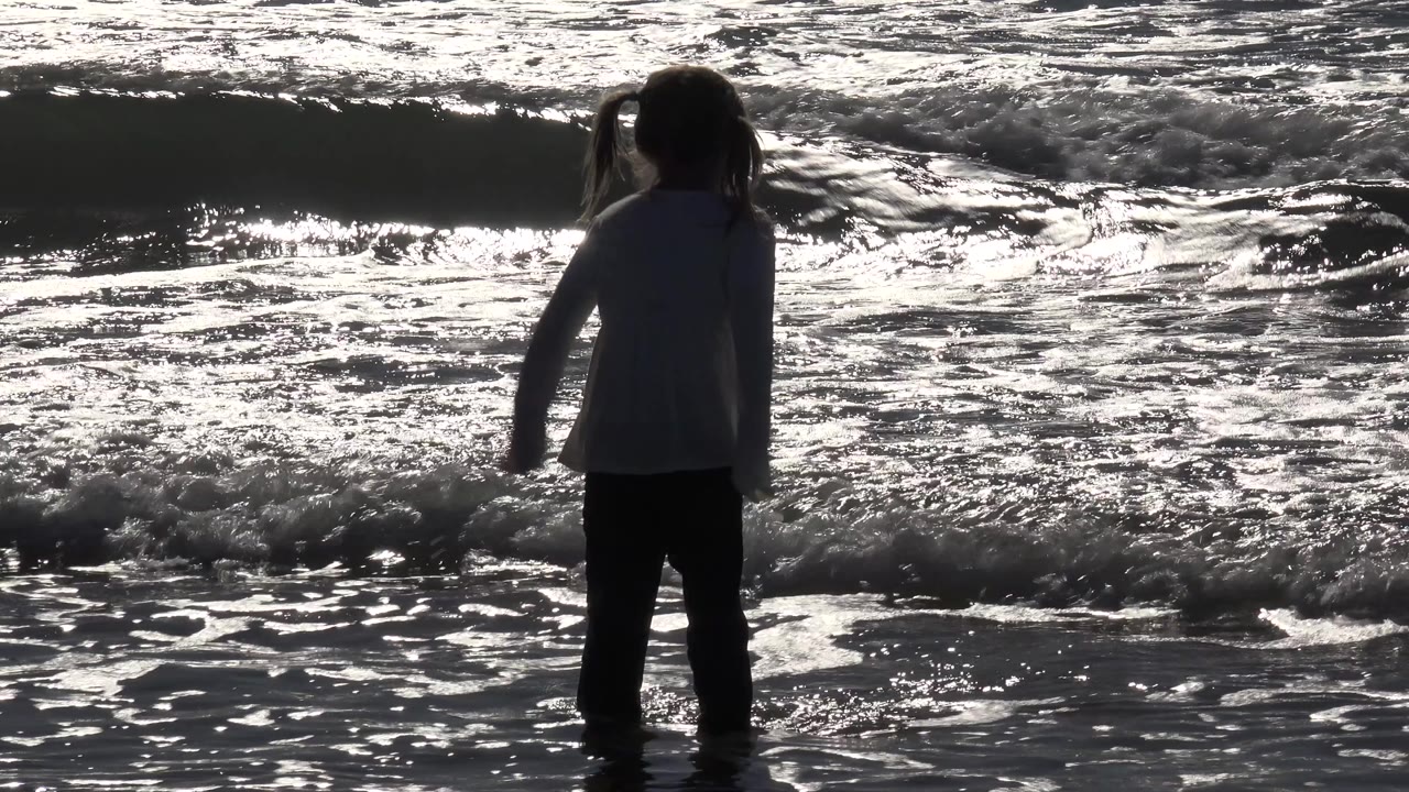 At Huntington Beach, California, the girl remains near the shoreline as a low wave curls and white foam crosses the foreground.