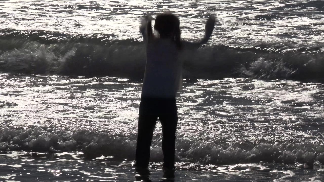 At Huntington Beach, California, the girl lifts both arms while a darker wave rises behind her in the backlit water.
