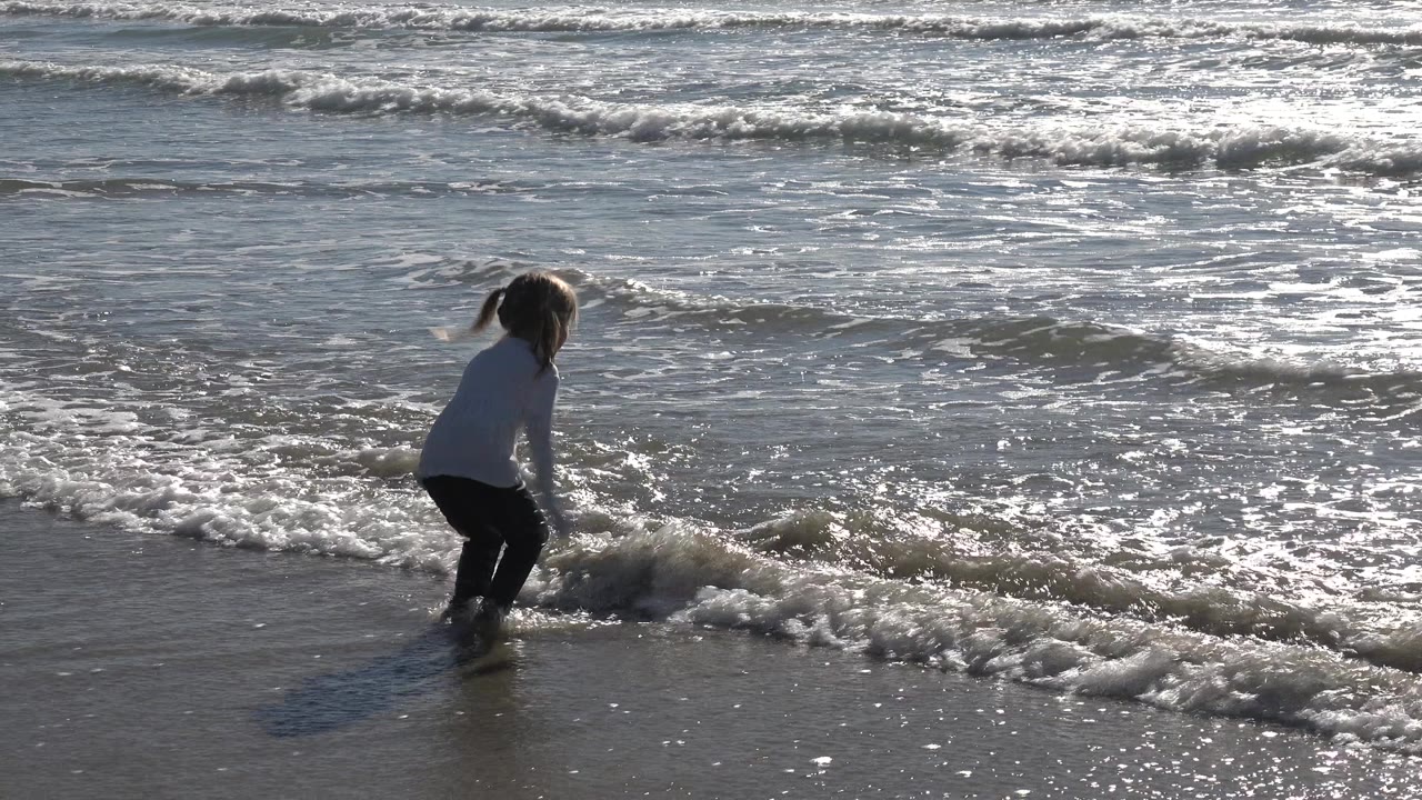 At Huntington Beach, California, a young girl in a light top and dark pants steps toward a small foamy wave as sun glare reflects across the surf.