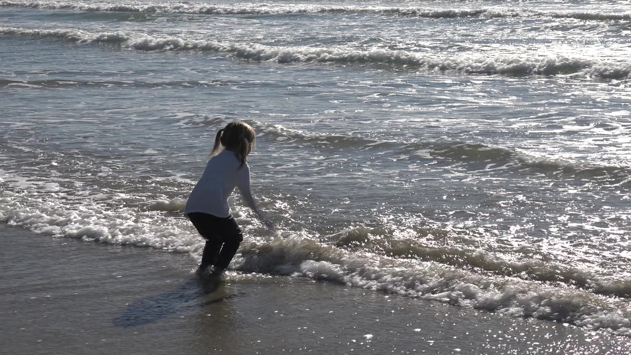 At Huntington Beach, California, the girl crouches slightly in ankle-deep foam while small waves and bright reflected water fill the background.