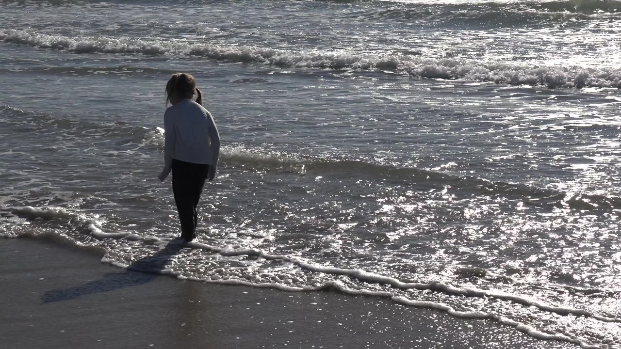 Young girl walks along the wet shoreline with foam around her feet.