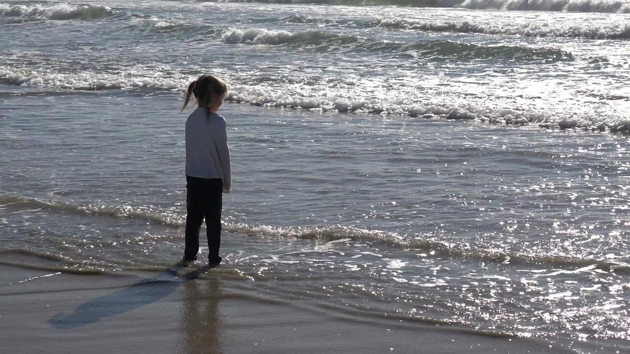 A young girl stands still at the Huntington Beach waterline as a thin wave reaches her feet and bright ocean glare fills the background.