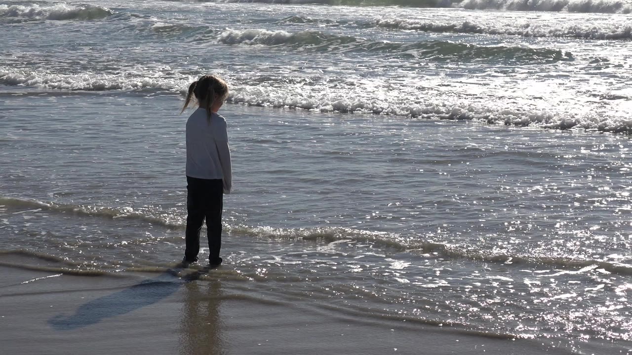 A young girl faces the Huntington Beach surf from wet sand while small foam lines approach and the sunlight reflects across the water.