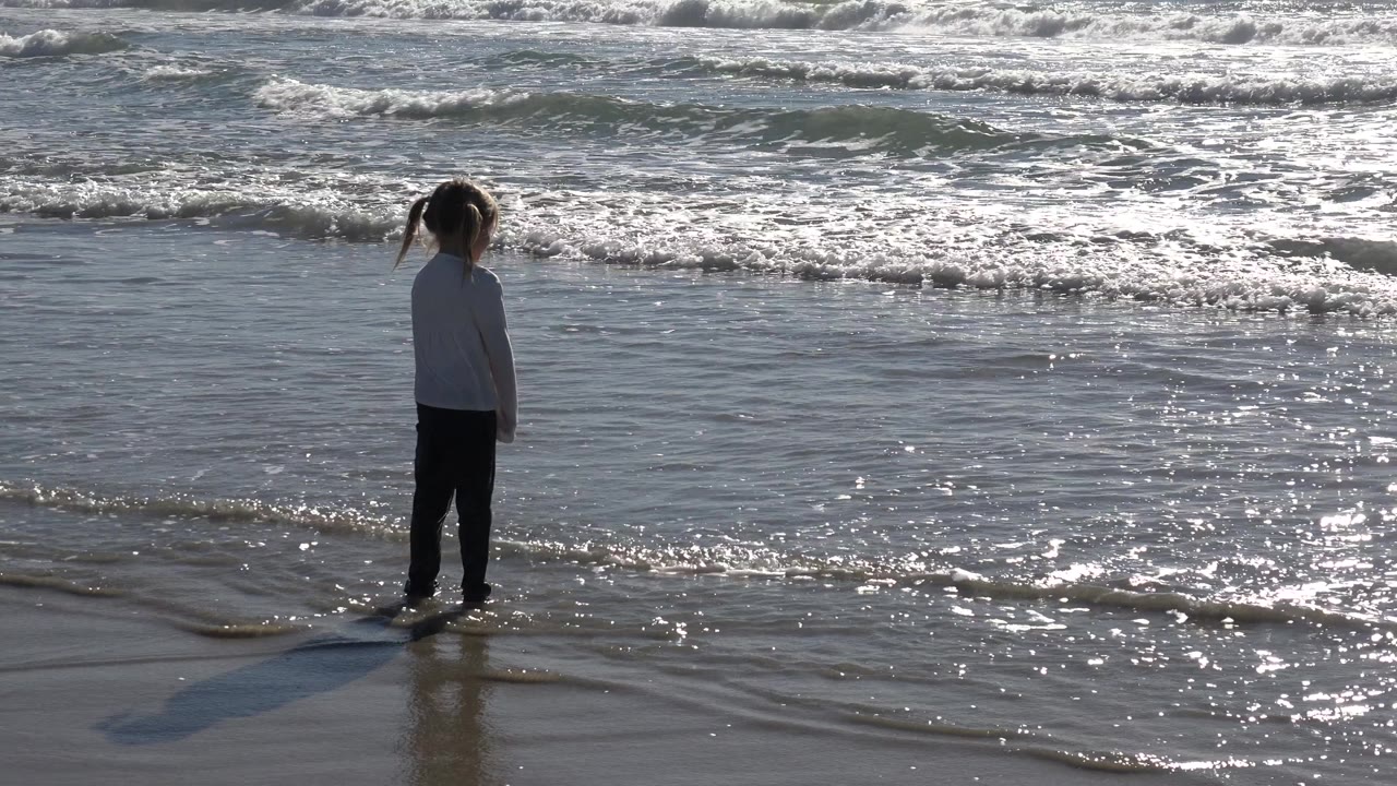 The girl remains near the shoreline at Huntington Beach with rippled shallow water around her feet and white surf bands across the background.