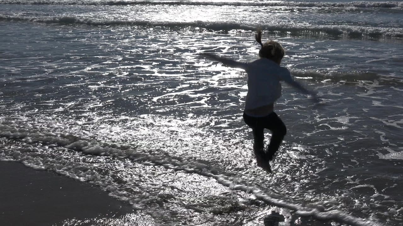 At Huntington Beach, California, a child jumps in shallow surf as bright reflected light spreads across the wet sand and incoming foam.