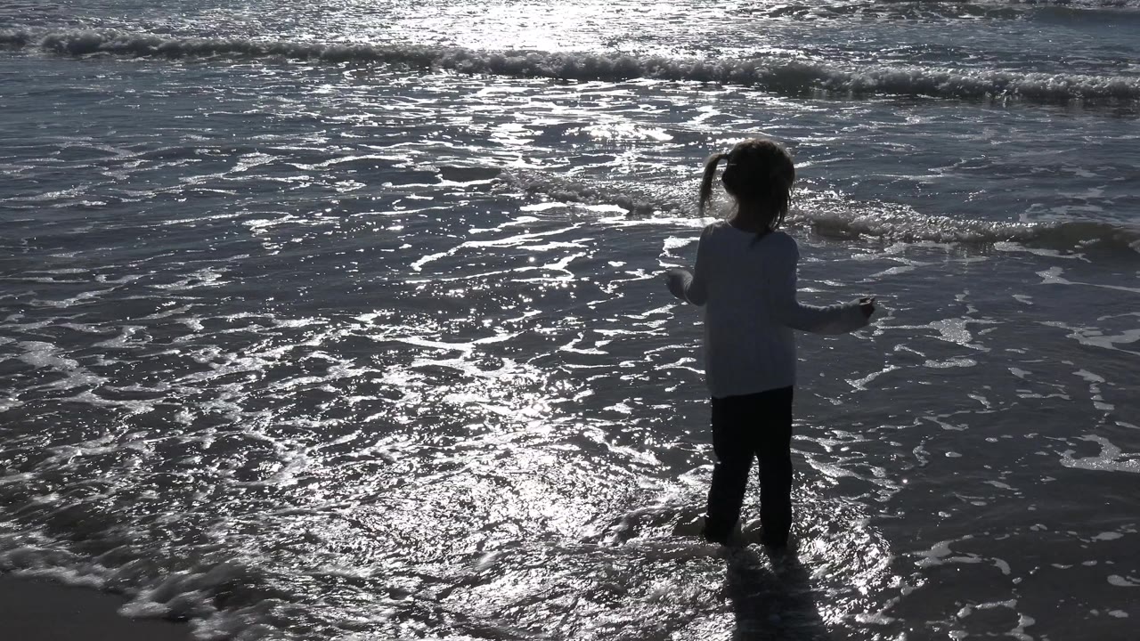 At Huntington Beach, California, the child stands in shallow water facing the ocean, with a foamy wave band and dark backlit water behind them.