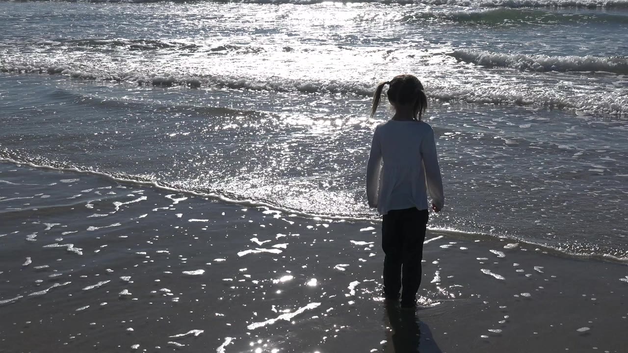 At Huntington Beach, a young girl faces the ocean from shallow water as low waves and bright reflections fill the background.