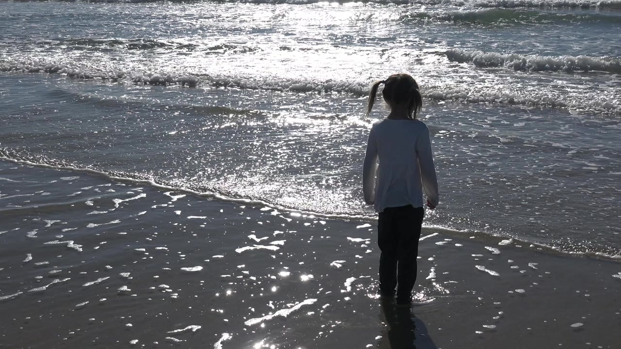 Young girl facing small waves on reflective wet sand.