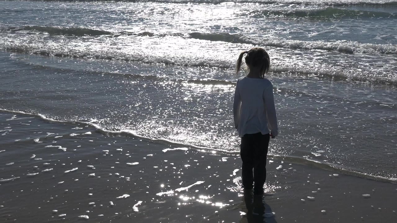 At Huntington Beach, the girl stands slightly farther right in the frame as foamy water and silver highlights spread across the ocean behind her.