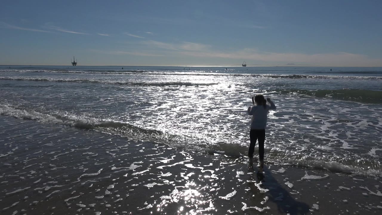 A young girl stands near the edge of small waves at Huntington Beach, California, with bright reflected sunlight stretching across the water behind her.