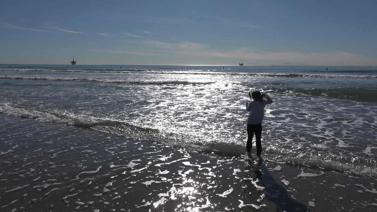 The girl faces the water at Huntington Beach, California, as a low wave line rolls toward her through bright reflective surf.