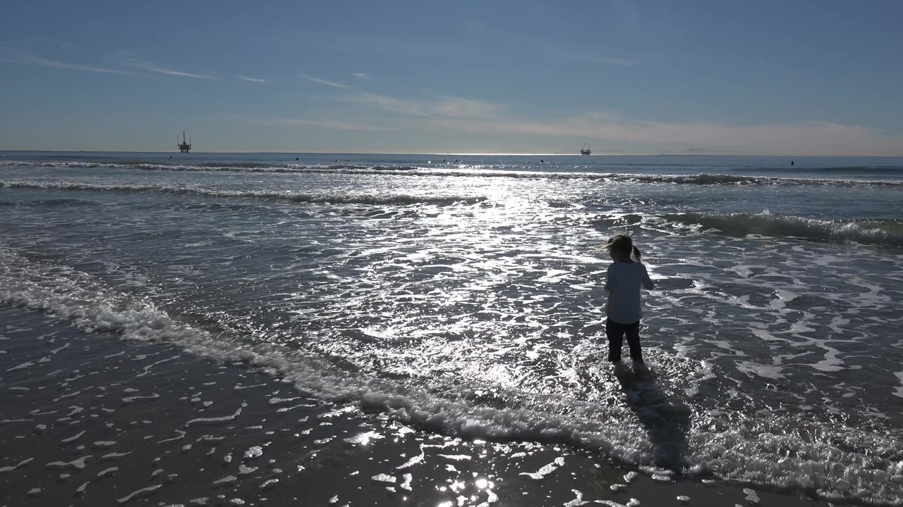 A young girl stands farther right in the frame at Huntington Beach, California, with wave foam spreading around her feet and intense light on the water.