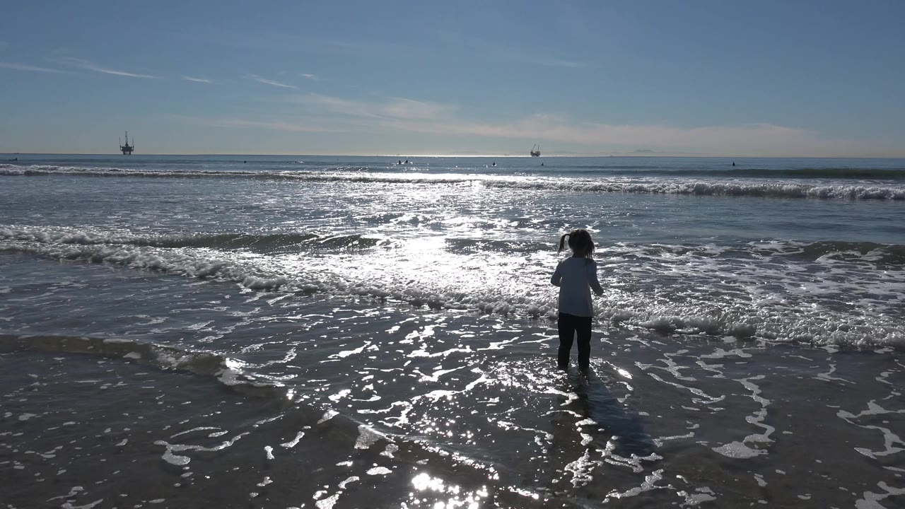 A young girl stands in foamy shallow water at Huntington Beach, California, with wet sand reflecting the sun and distant offshore structures near the horizon.