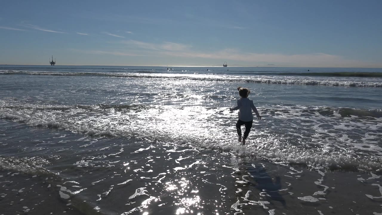 A young girl jumps through the shallow Huntington Beach shorebreak near frame right, while sunlit foam and small waves fill the foreground and midground.