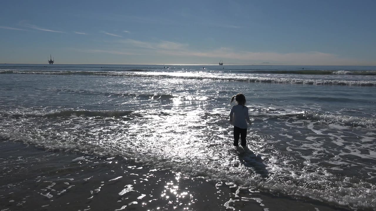 Young girl standing in shallow ocean water with a long shadow on wet sand.