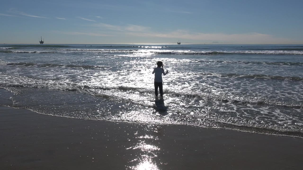 A child stands near the Huntington Beach shoreline in shallow foamy water, with sun glare on the ocean and offshore platforms on the horizon.