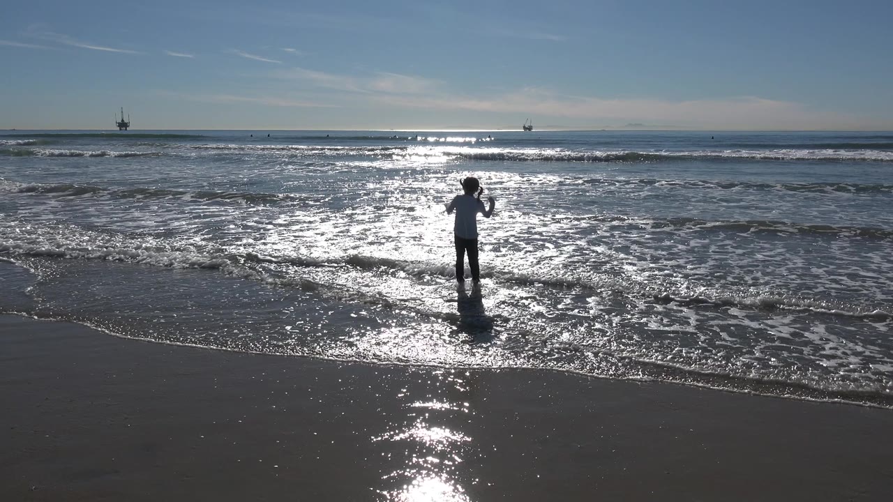 The child raises both arms in the shallow Huntington Beach surf while bright reflected sunlight spreads across the water behind them.