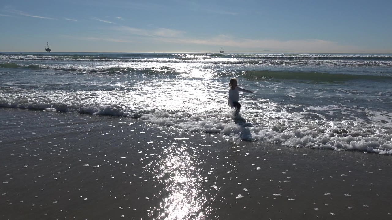 At Huntington Beach, California, a young girl stands near the breaking wave line while bright sunlight reflects across wet sand and the ocean surface.
