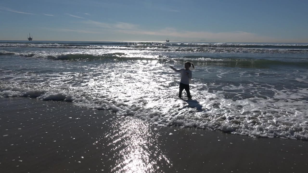 At Huntington Beach, California, the child raises her arms near the waterline as white surf rolls in and distant offshore structures sit on the horizon.