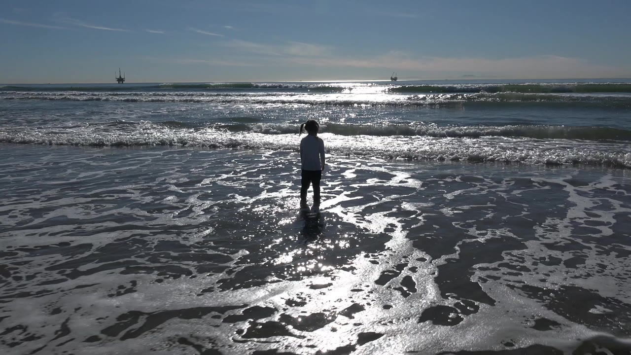 A child stands near the center of the frame in shallow water at Huntington Beach, California, facing bright backlit waves under a clear sky.