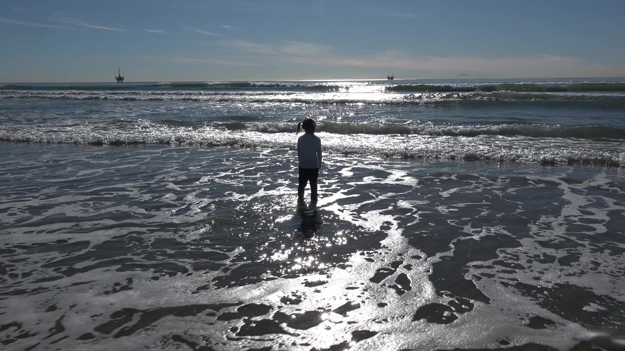 Child facing the ocean as foam spreads over wet sand