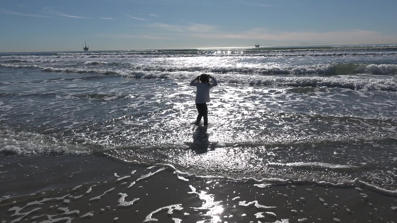 A young girl stands at the Huntington Beach shoreline with both hands near her head while bright surf glare spreads across the wet sand.