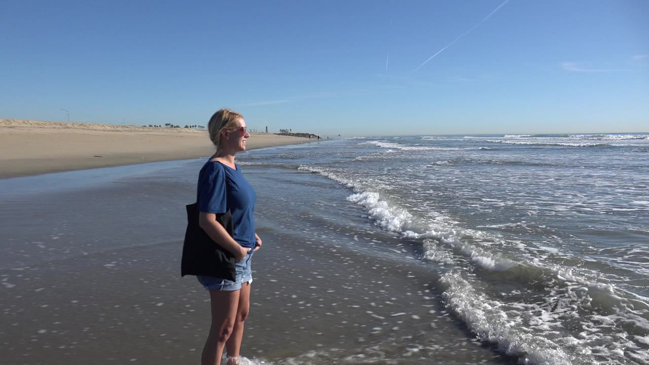 A blonde woman in sunglasses stands near the waterline at Huntington Beach as a small wave runs up the wet sand beside her.