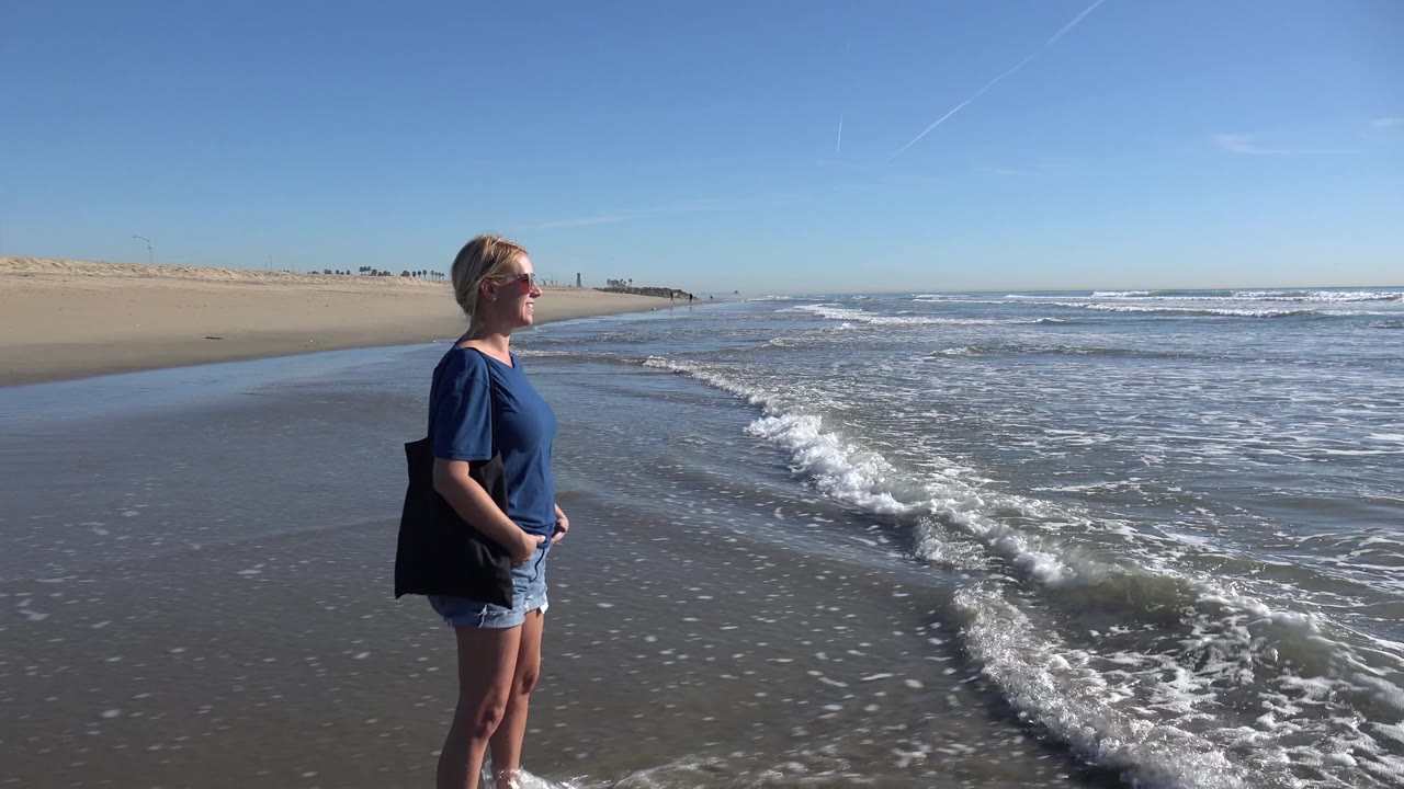 Woman in blue shirt facing ocean while waves wash along the shore.