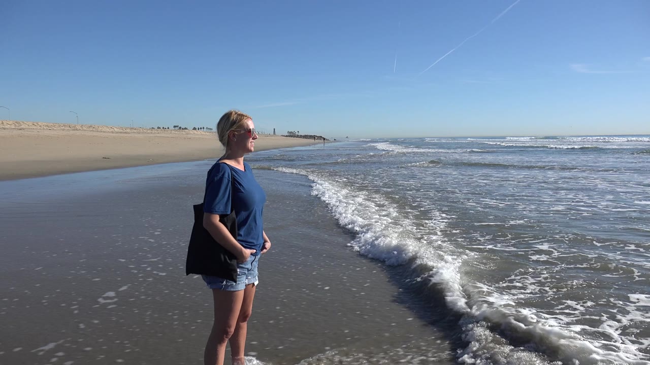 A larger foamy wave reaches closer to the woman at Huntington Beach, with her standing near the left side and the ocean spread across the right.