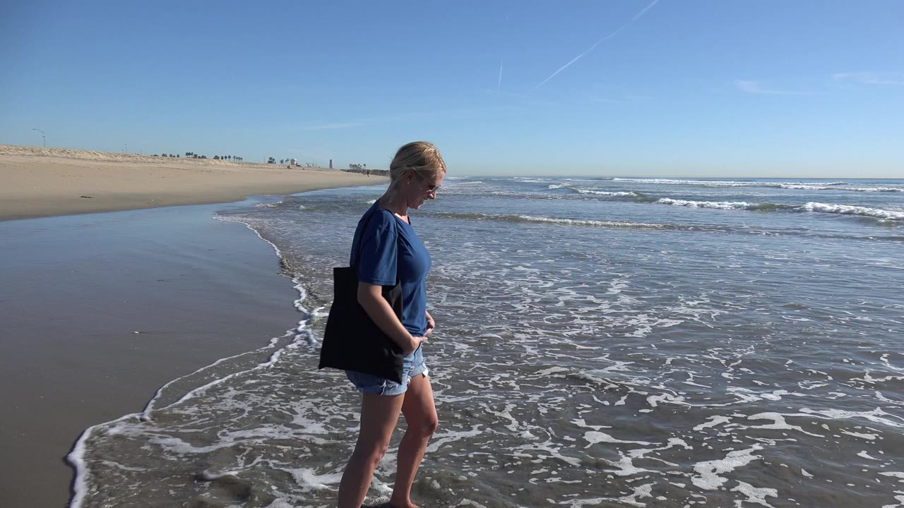 A blonde woman in a blue shirt walks barefoot along wet sand beside the incoming surf at Huntington Beach, California, with open beach stretching behind her.