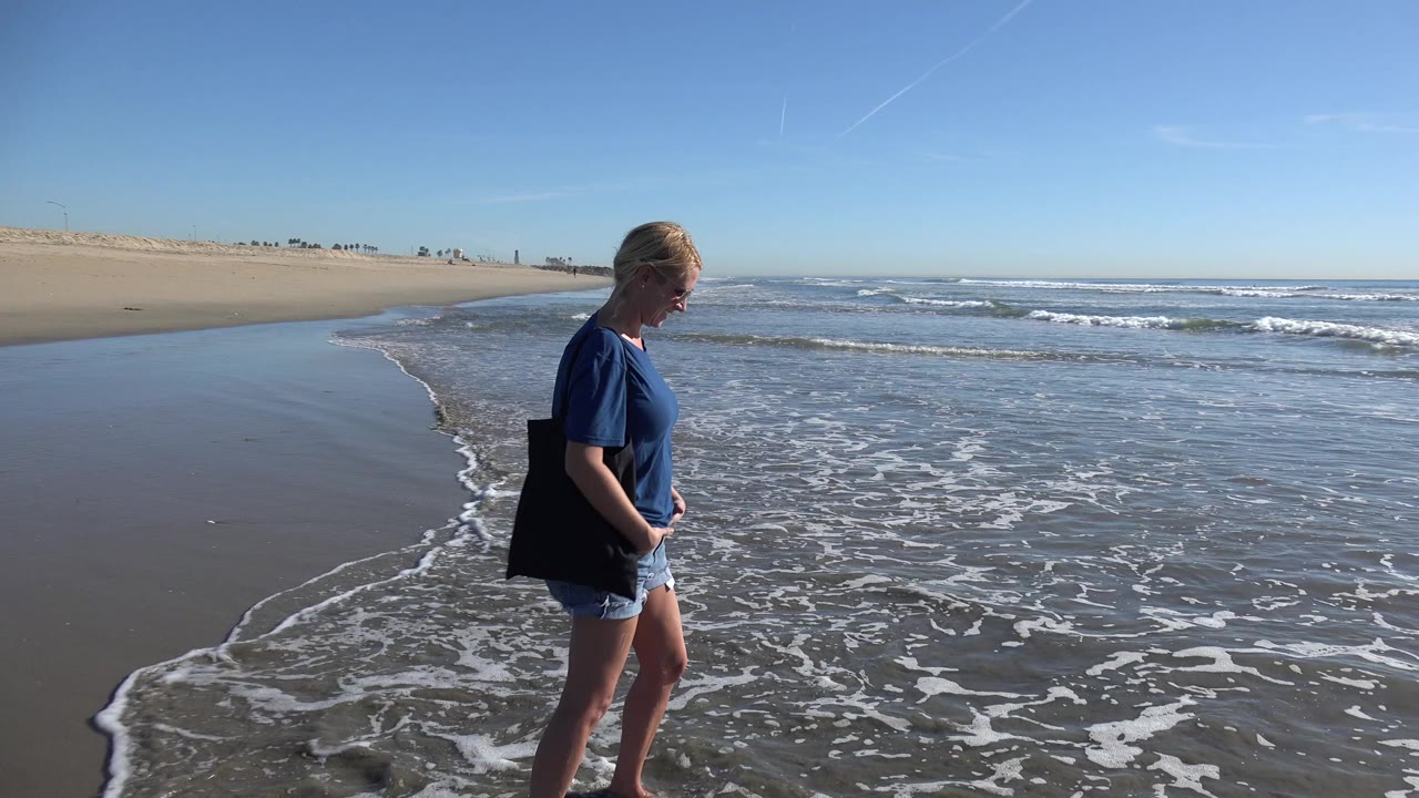 Woman in blue walking along wet shoreline with waves beside her.