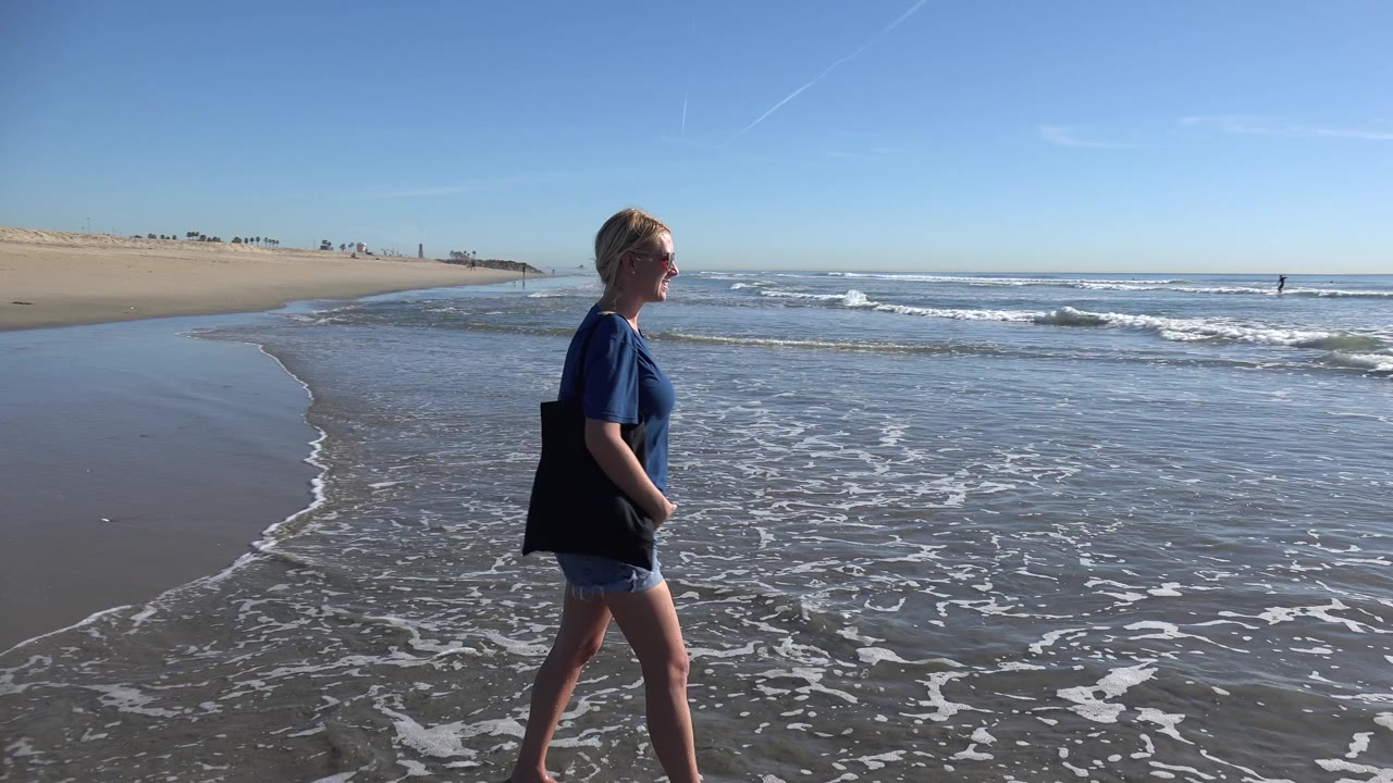 At Huntington Beach, California, the woman moves farther across the frame near shallow foam while the ocean horizon and a broad empty sandbar remain visible behind her.