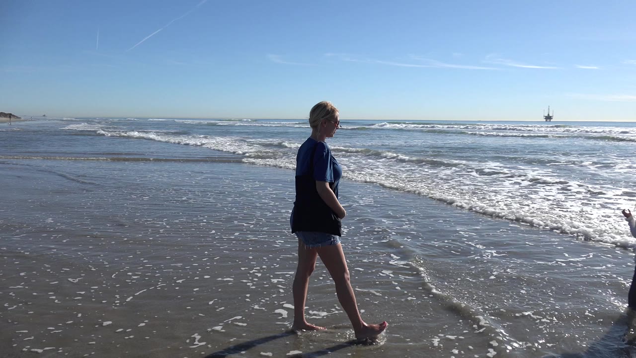 At Huntington Beach, California, a woman in a dark shirt walks barefoot along wet sand as white surf rolls in behind her.