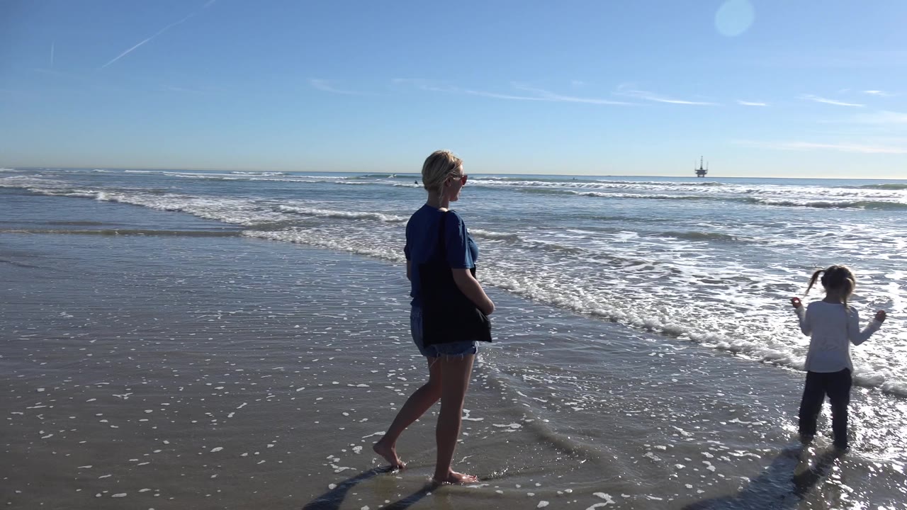 At Huntington Beach, California, a woman stands near the surf while a young child appears at the right edge beside foamy shallow water.
