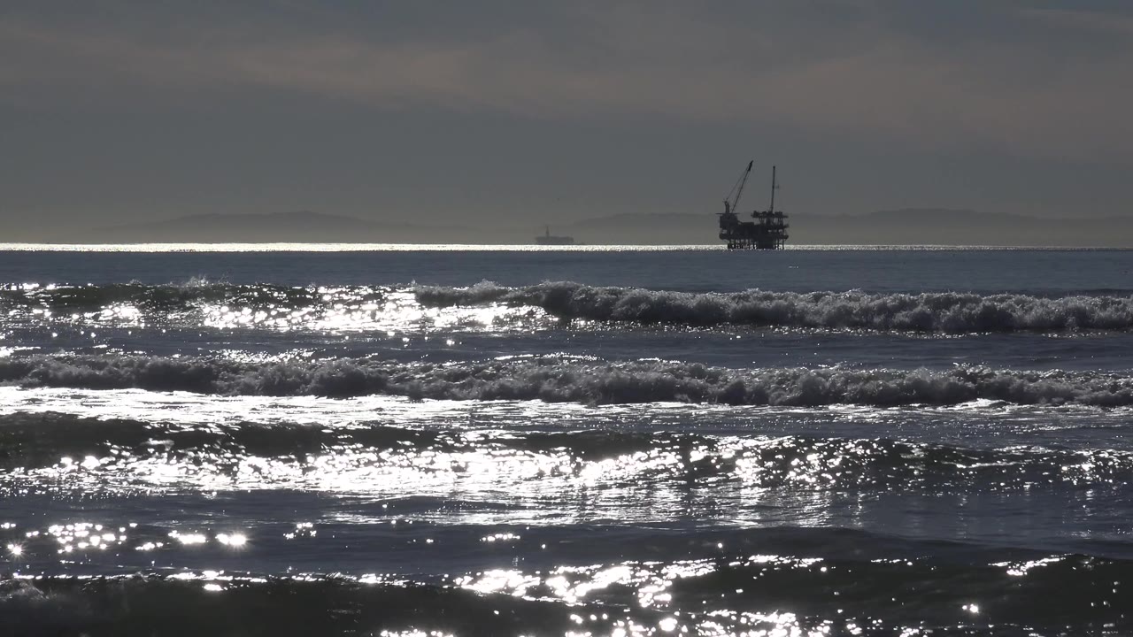 A Huntington Beach surf line breaks across the midground as silver highlights scatter over wet water and a small offshore structure or vessel remains near the horizon, with no visible people.