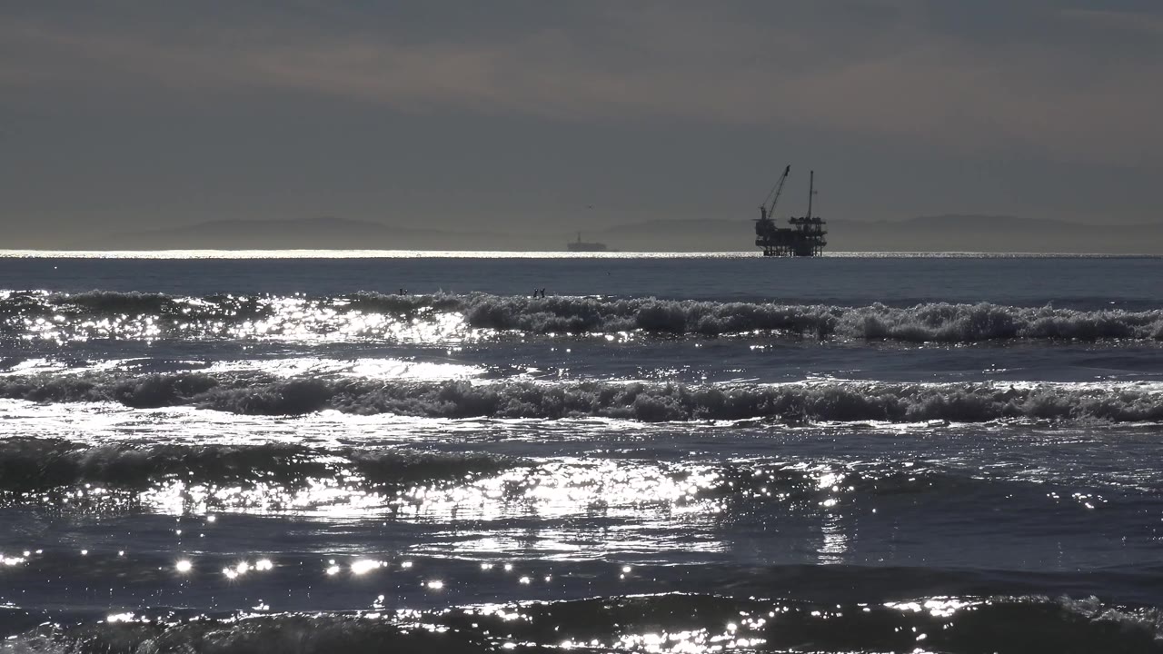Sunlight flashes across Huntington Beach waves while the distant offshore silhouette shifts toward the right side of the composition and no visible people appear in the surf.