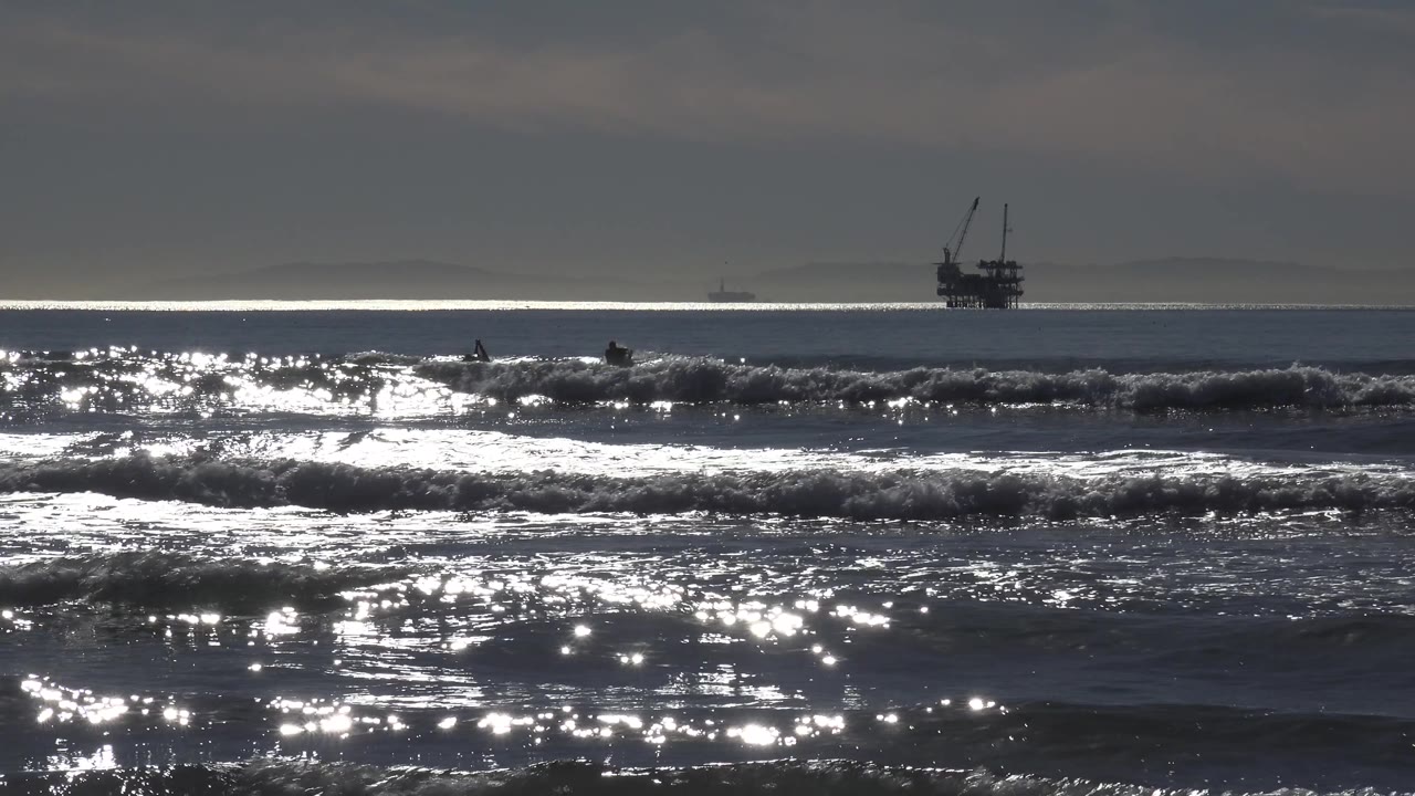 Choppy Huntington Beach surf rolls through the foreground and midground as a distant offshore structure sits left of center under a subdued sky, with no visible people.