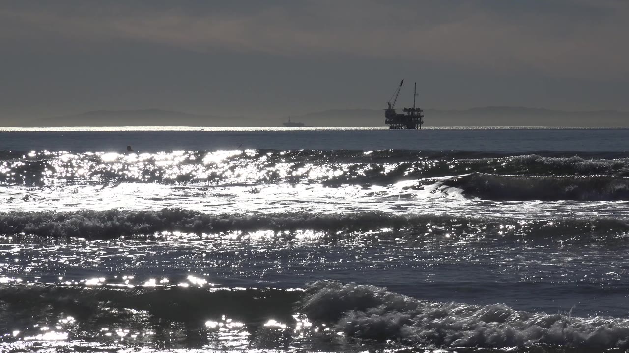At Huntington Beach, the distant ship appears near the center horizon as sun glare spreads across the water and low surf breaks in front, with no visible people.