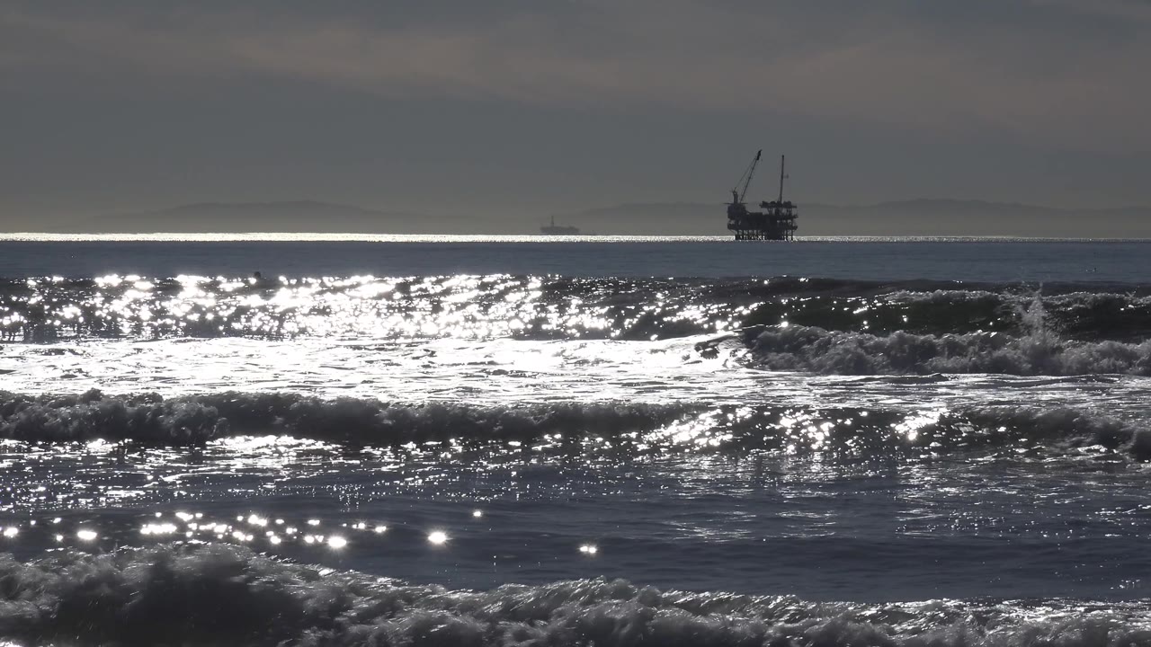 At Huntington Beach, the ship sits toward the right side of the horizon while choppy waves and silver water reflections fill the foreground, with no visible people.