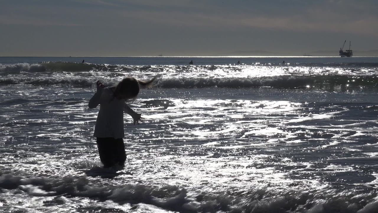A young girl in Huntington Beach stands in foamy shallow water, silhouetted against bright reflected ocean light and distant offshore boats.